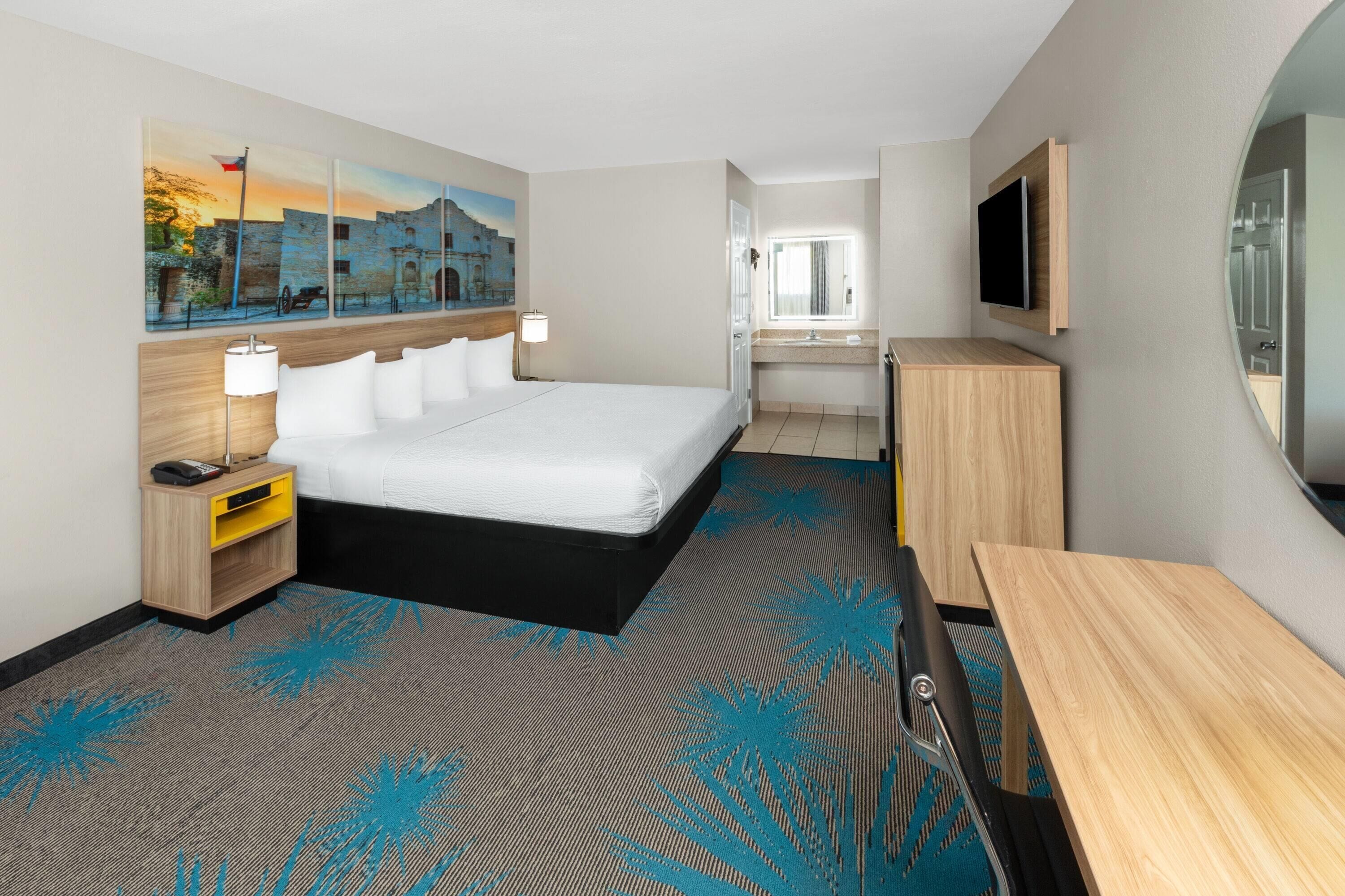 A hotel room with a bed, bedside table, lamp, TV, and a painting on the wall.