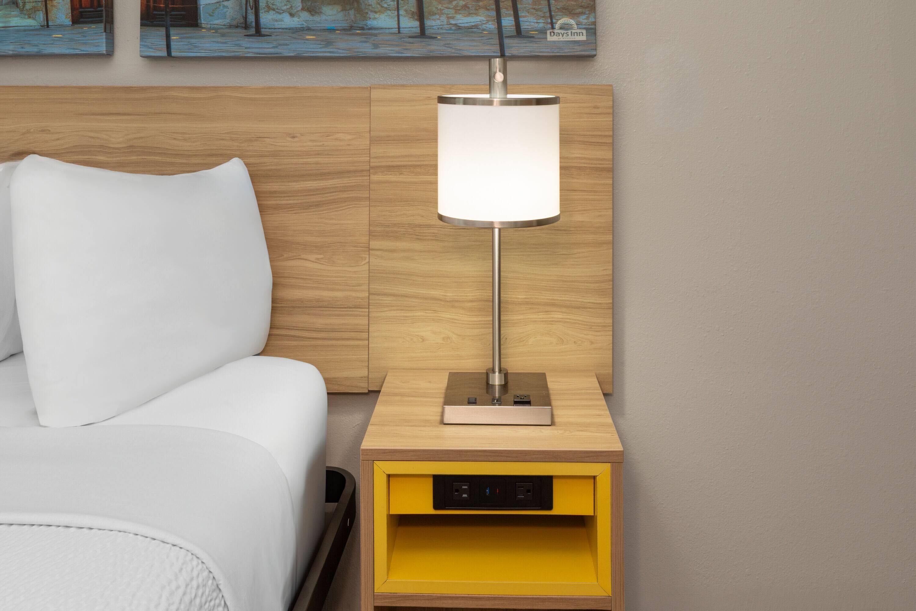 Habitación de hotel con cabecera de madera, cama blanca, mesita de noche con lámpara y cajón amarillo.