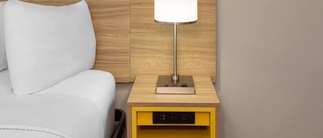 Habitación de hotel con cabecera de madera, cama blanca, mesita de noche con lámpara y cajón amarillo.