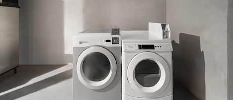 A laundry area with two front-loading washers and dryers, a small shelf with a container, and a vent on the left.