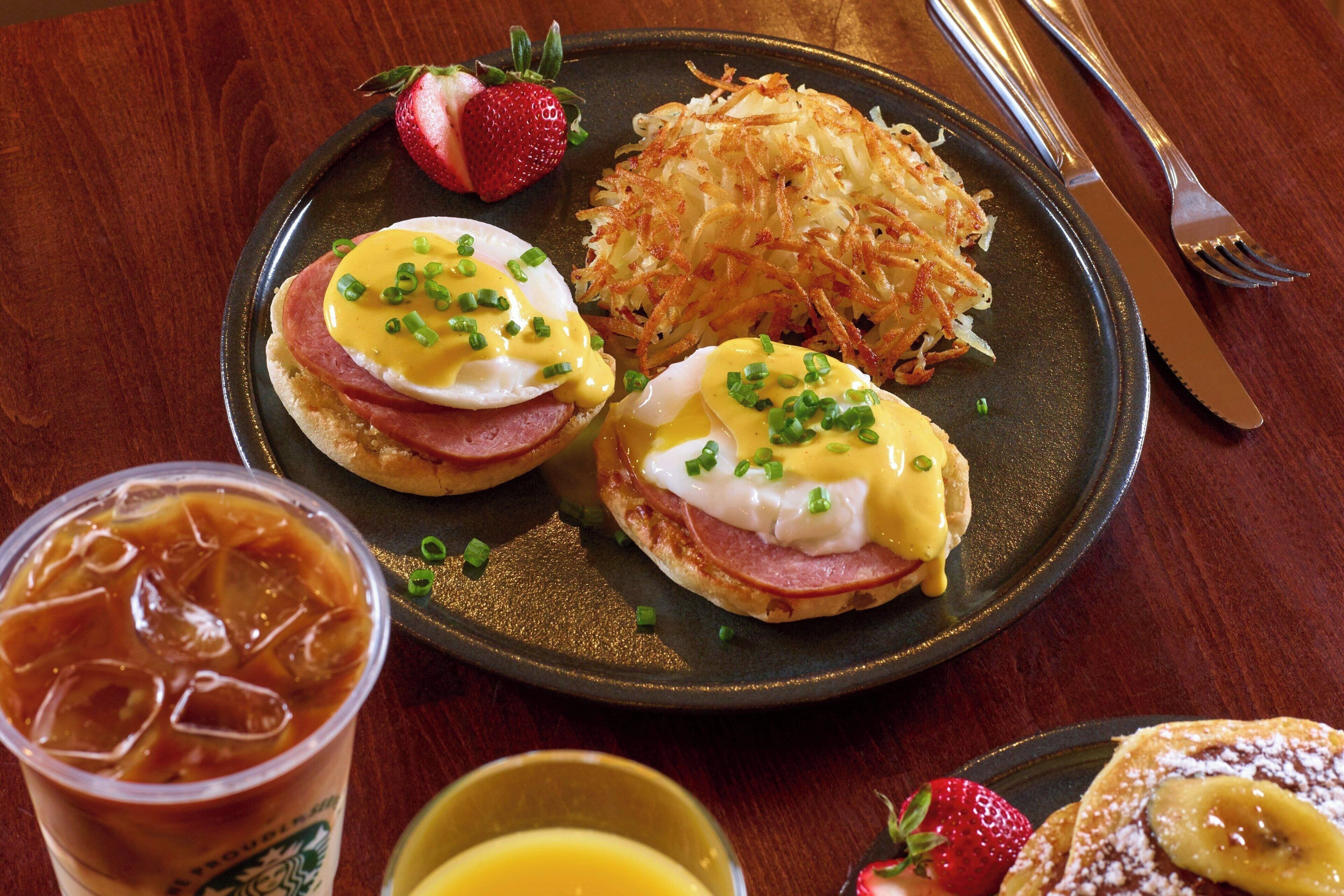 A plate with eggs Benedict and hash browns, a glass of iced coffee, and a slice of banana bread with strawberries.