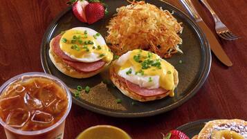 A plate with eggs Benedict and hash browns, a glass of iced coffee, and a slice of banana bread with strawberries.
