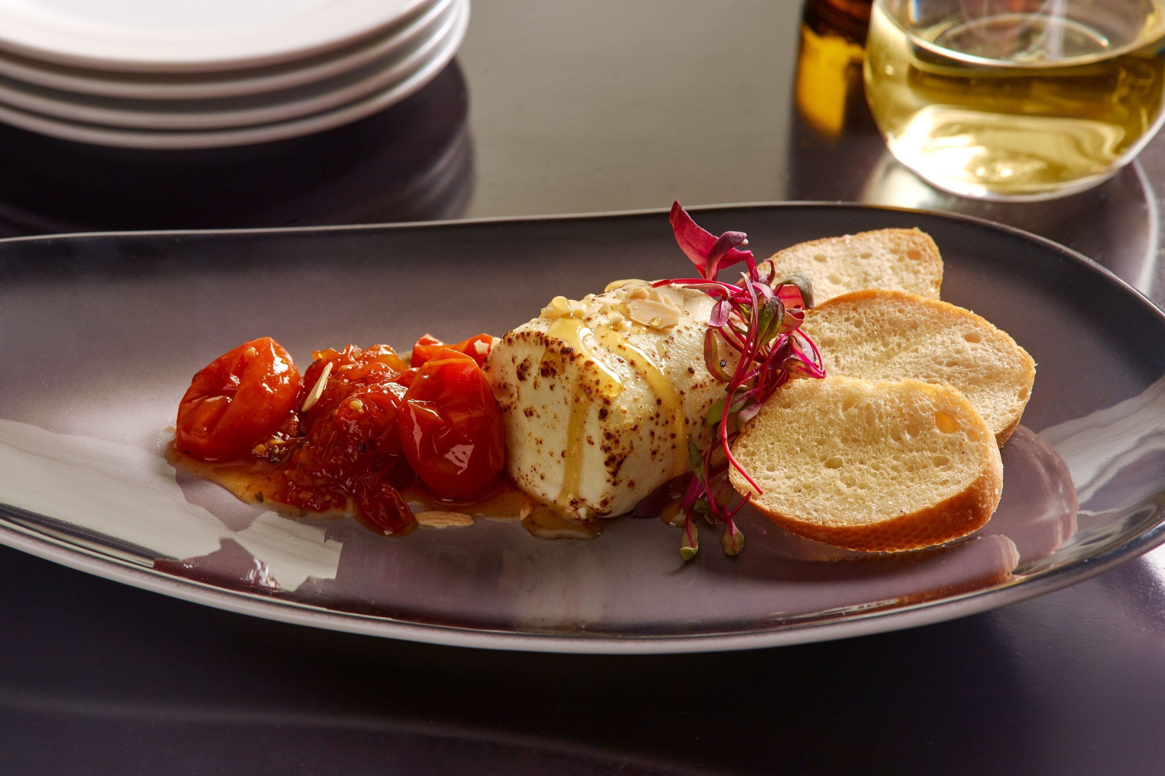 A plate of food with a white fish fillet, cherry tomatoes, and bread on a dark plate.