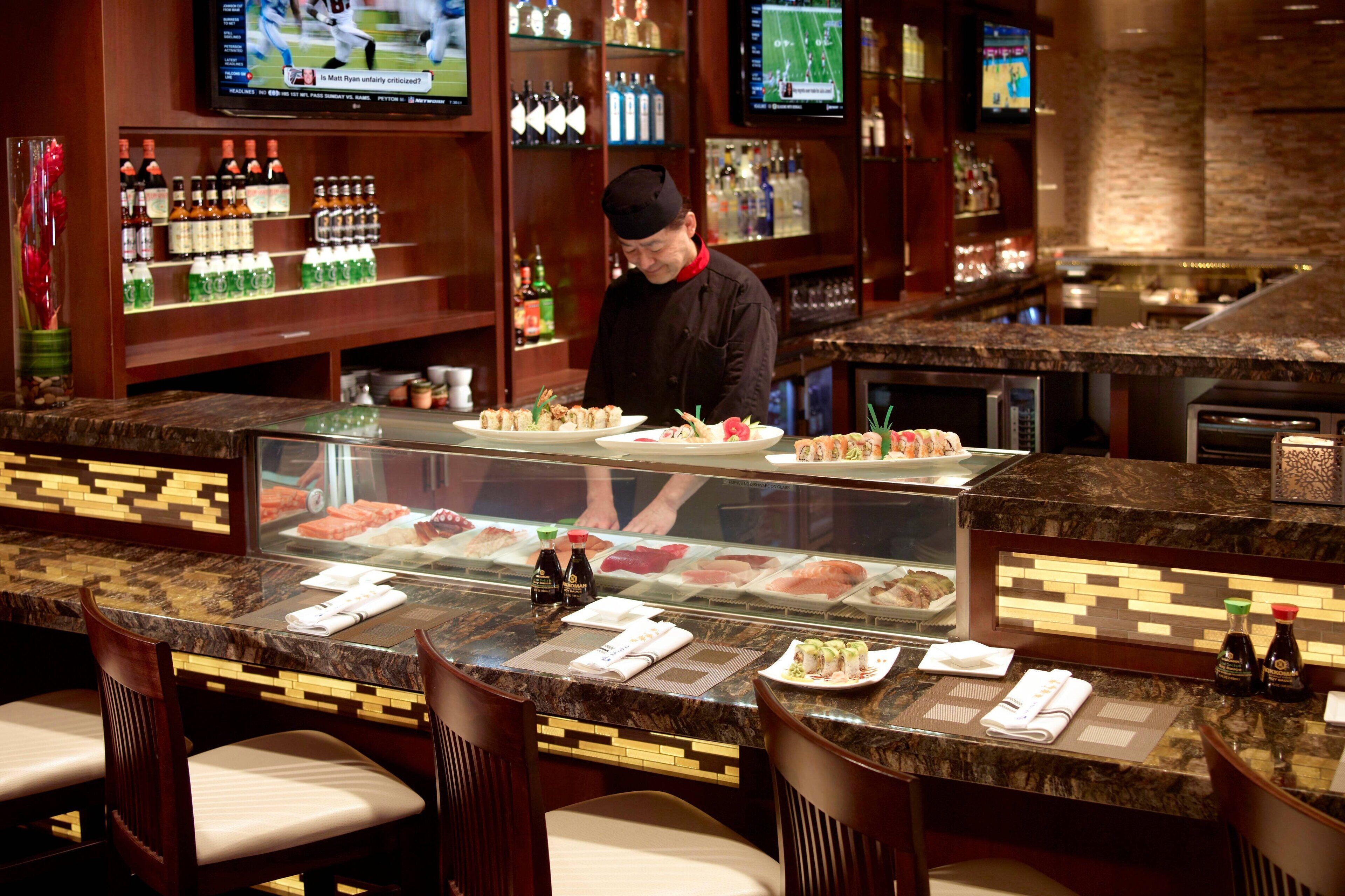 Un bar de sushi amb un xef preparant plats, una selecció de botelles a les prestatgeries i un taulell amb plats de sushi.