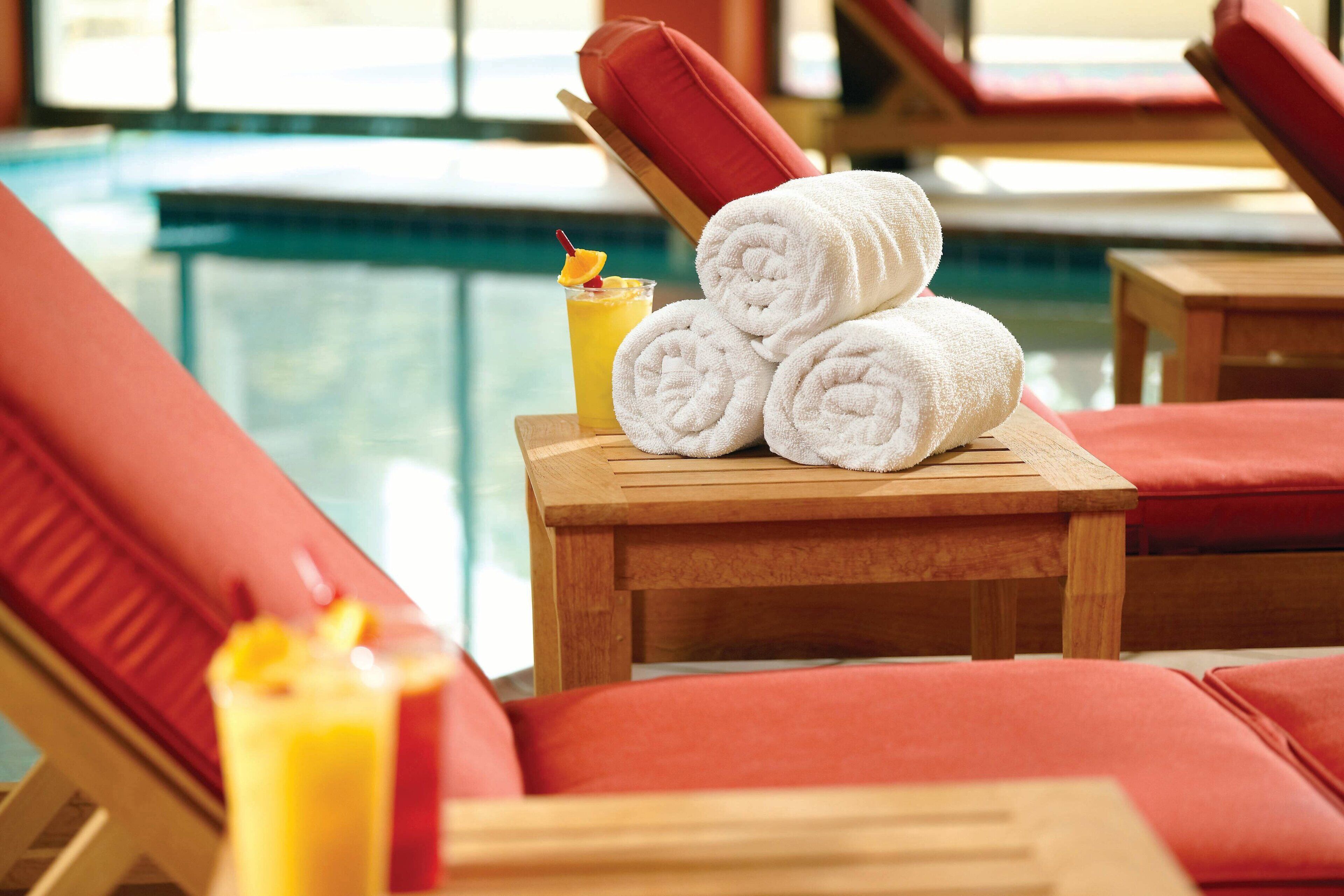 Un espace aménagé au bord de la piscine, avec des chaises longues, une table en bois sur laquelle sont posées des serviettes, et un verre de jus d’orange avec une paille.
