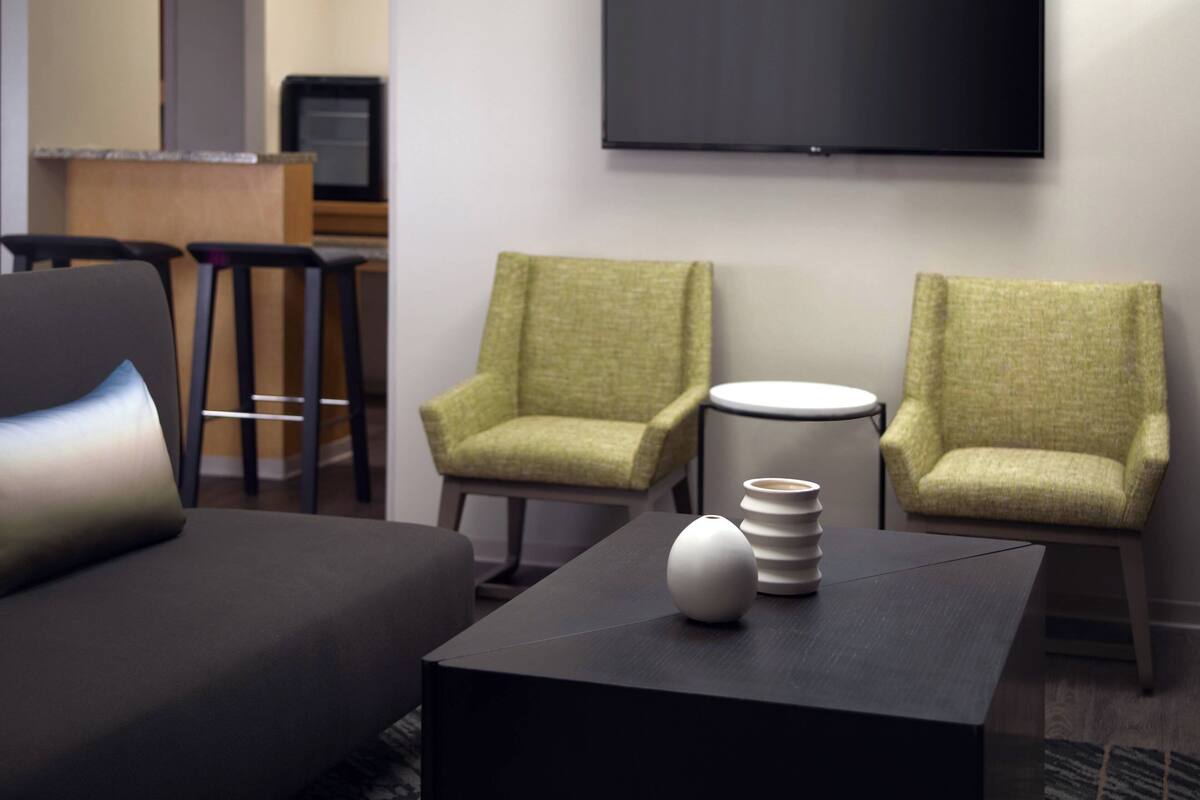Una habitación de hotel moderna con un sofá, dos sillones, una mesa de centro y un televisor de pantalla plana montado en la pared.