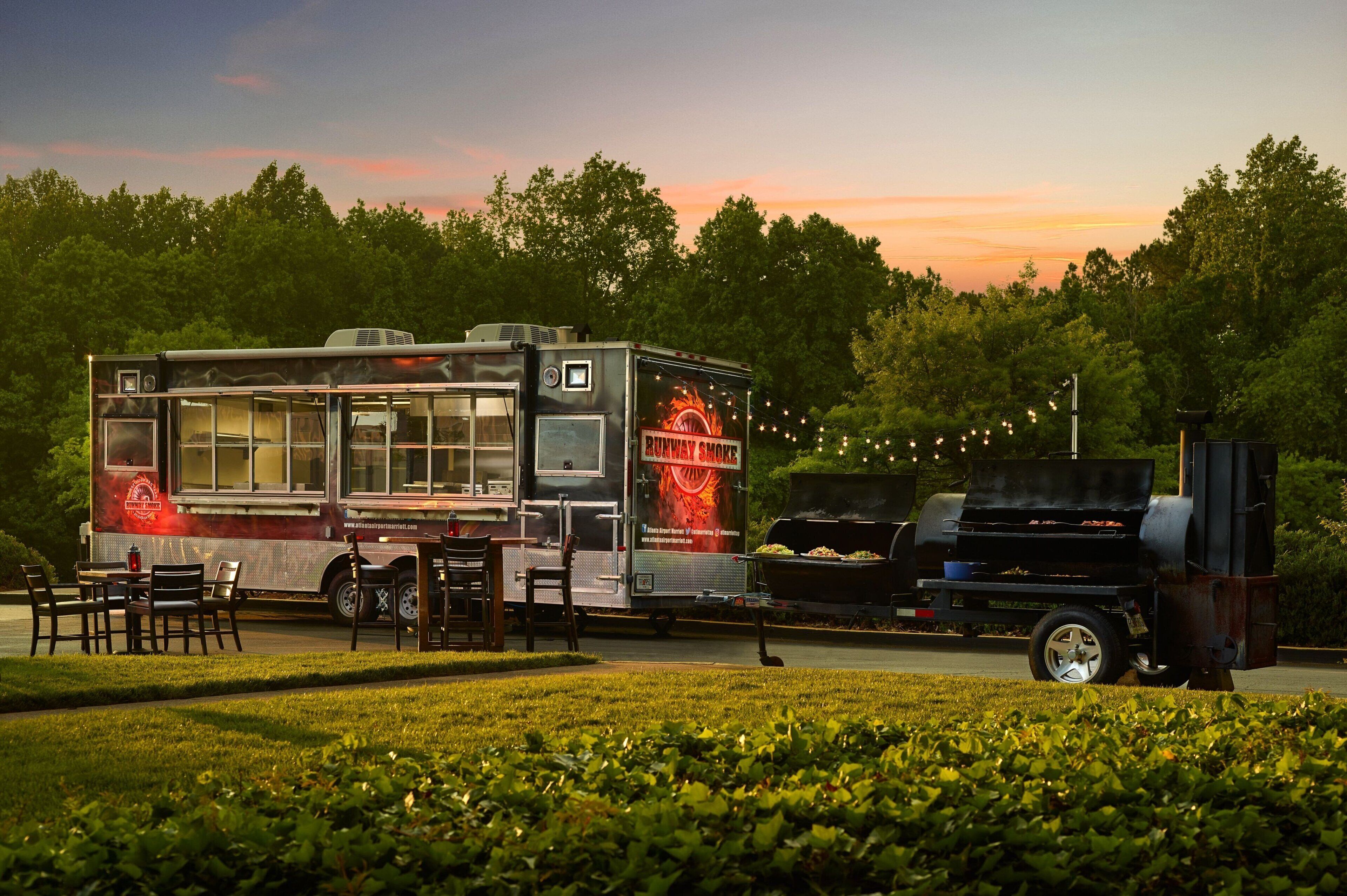 Un food truck amb una graella i zona de seients, davant d'un fons de trees i un capvespre.