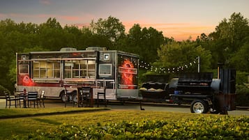 Un food truck amb una graella i zona de seients, davant d'un fons de trees i un capvespre.