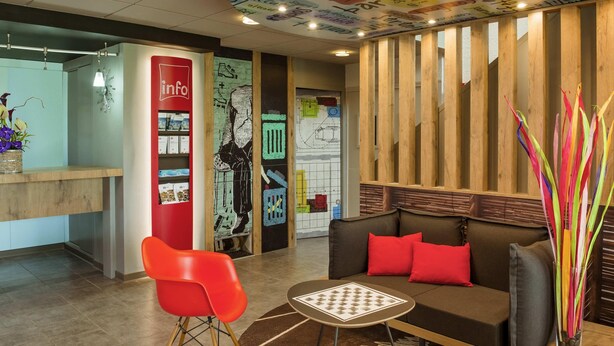 Un área de recepción con un mostrador de información rojo, un mural en blanco y negro, un mostrador de madera, un sofá marrón con cojines rojos y un jarrón de cristal con palitos de colores.