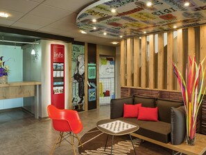 A reception area with a red information desk, a black and white mural, a wooden reception desk, a brown sofa with red cushions, and a glass vase with colorful sticks. - ibis Lyon Sud Saint Rambert d'Albon (Saint-Rambert-d'Albon)