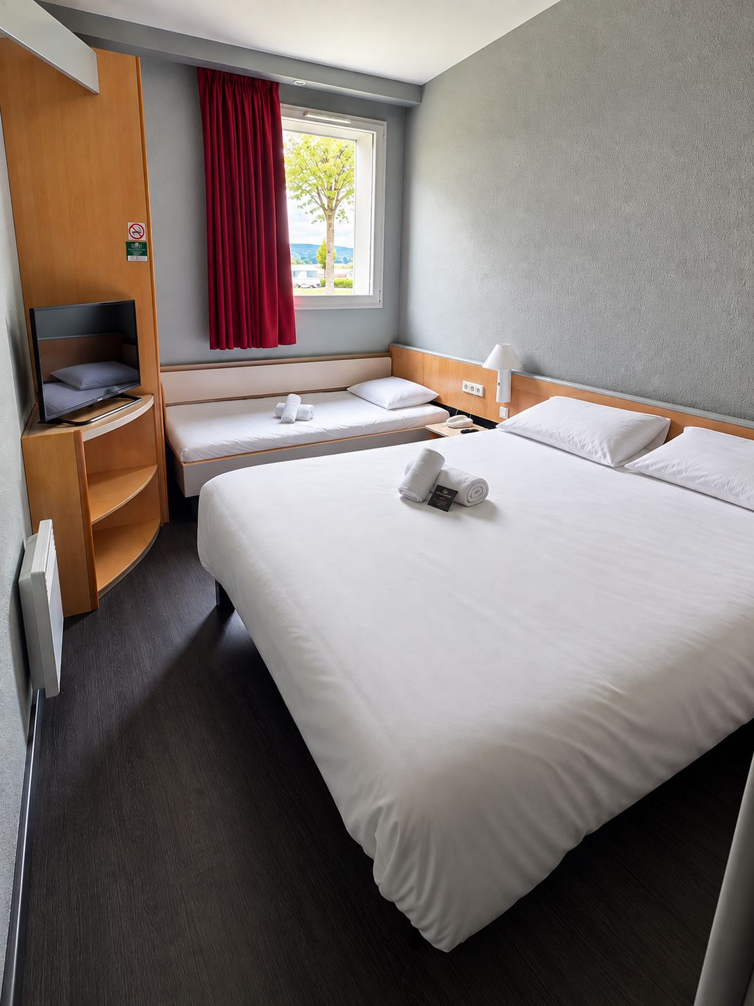 A hotel room with a bed, a TV, a desk, and a window with red curtains.