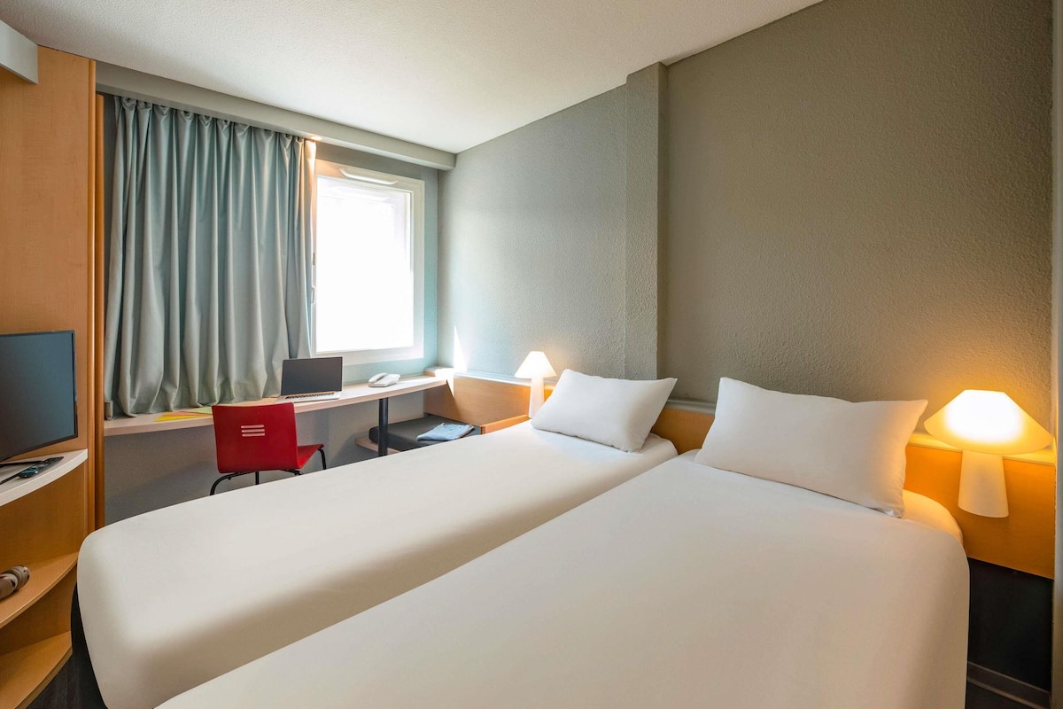 A hotel room with two beds, a desk with a laptop, a chair, a TV, and a window with curtains.