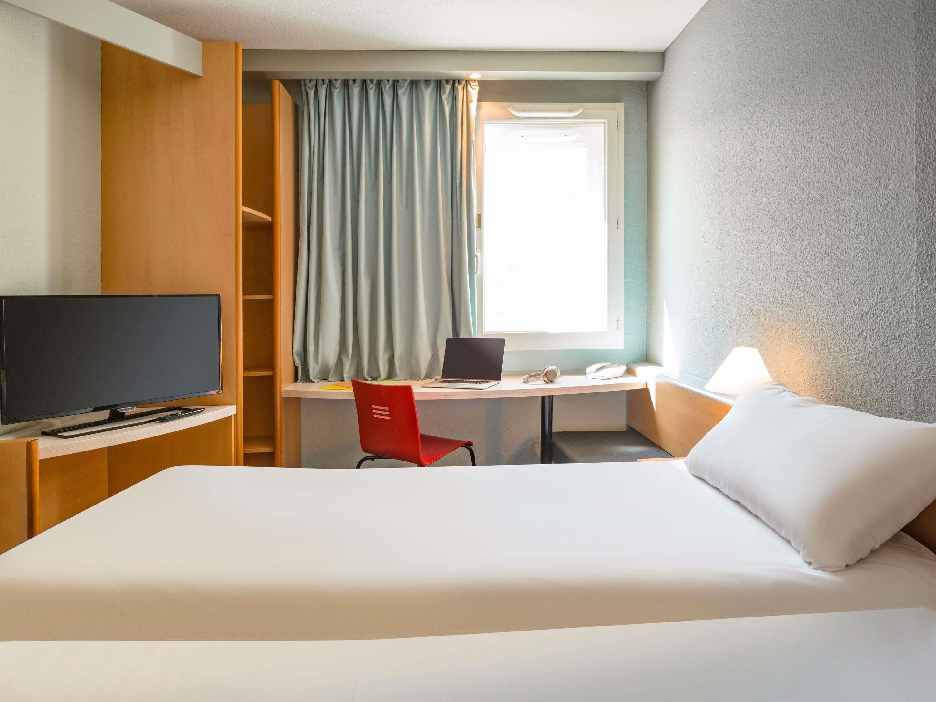 Una camera d'albergo con un letto, una scrivania con un portatile, una televisione e una finestra con tende.