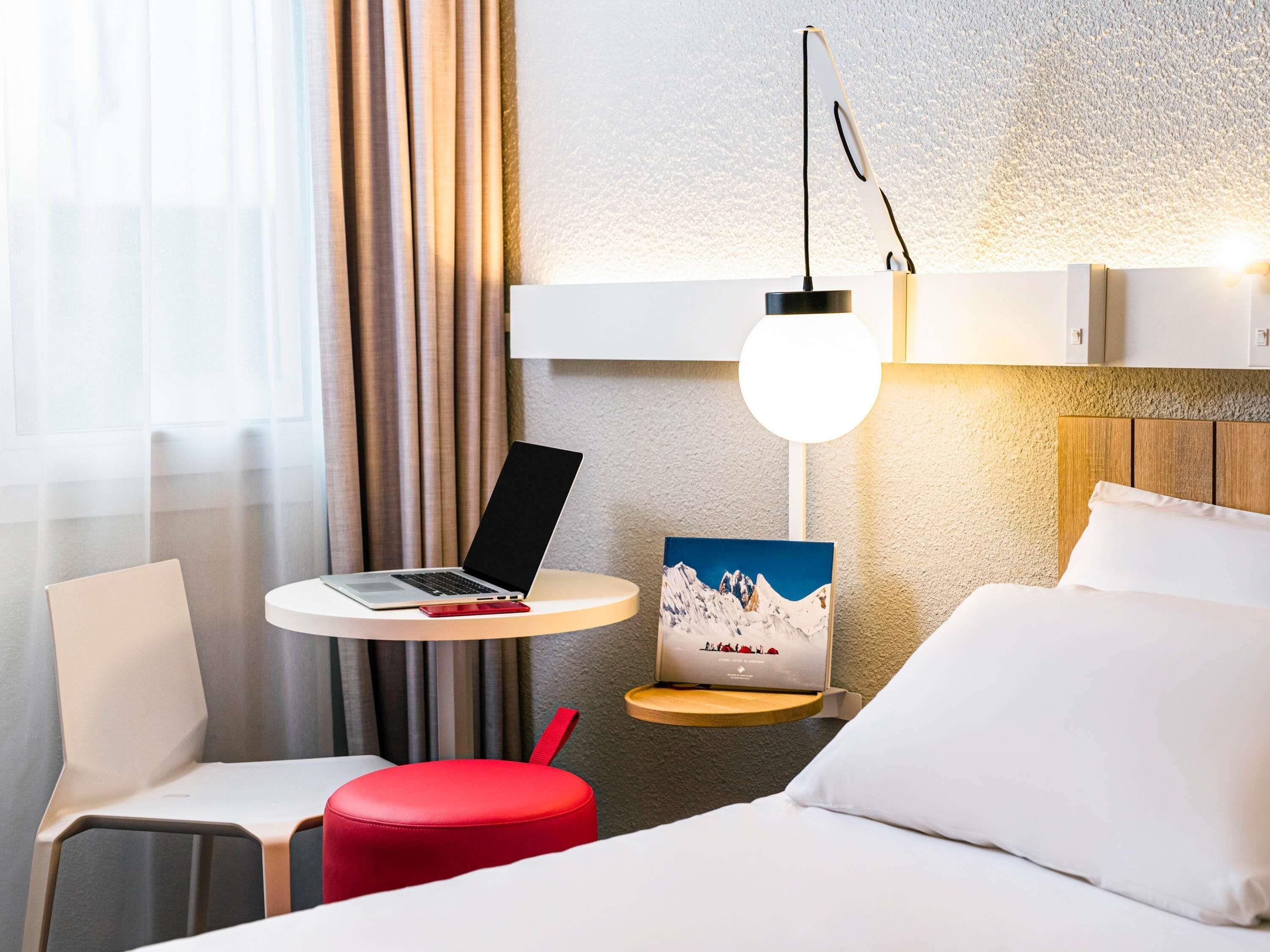 Habitación de hotel con cama, escritorio con computadora portátil, una otomana roja, un cuadro de montañas y una lámpara de pared.