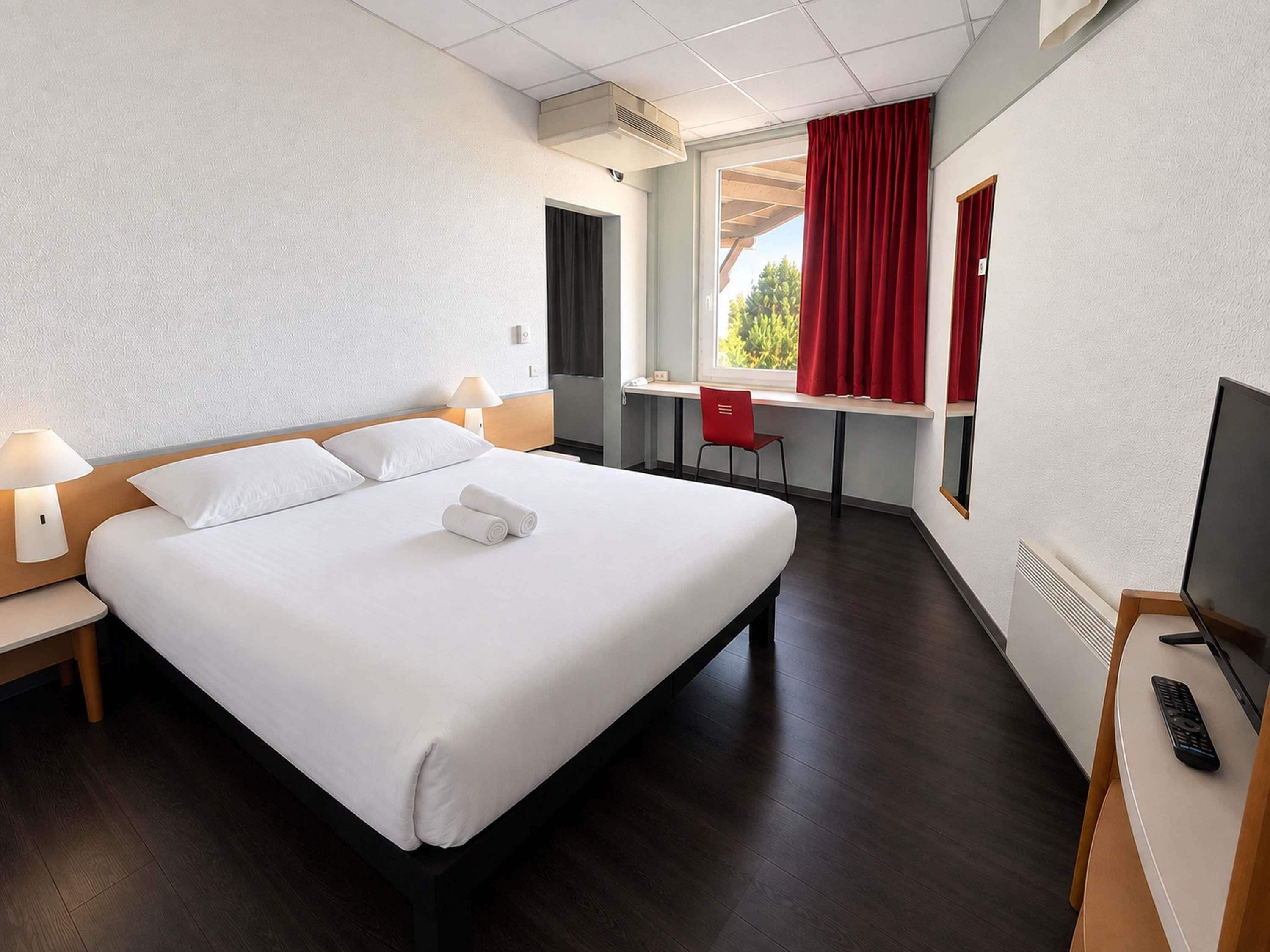 A hotel room with a bed, a desk, a chair, a TV, and a window with curtains.