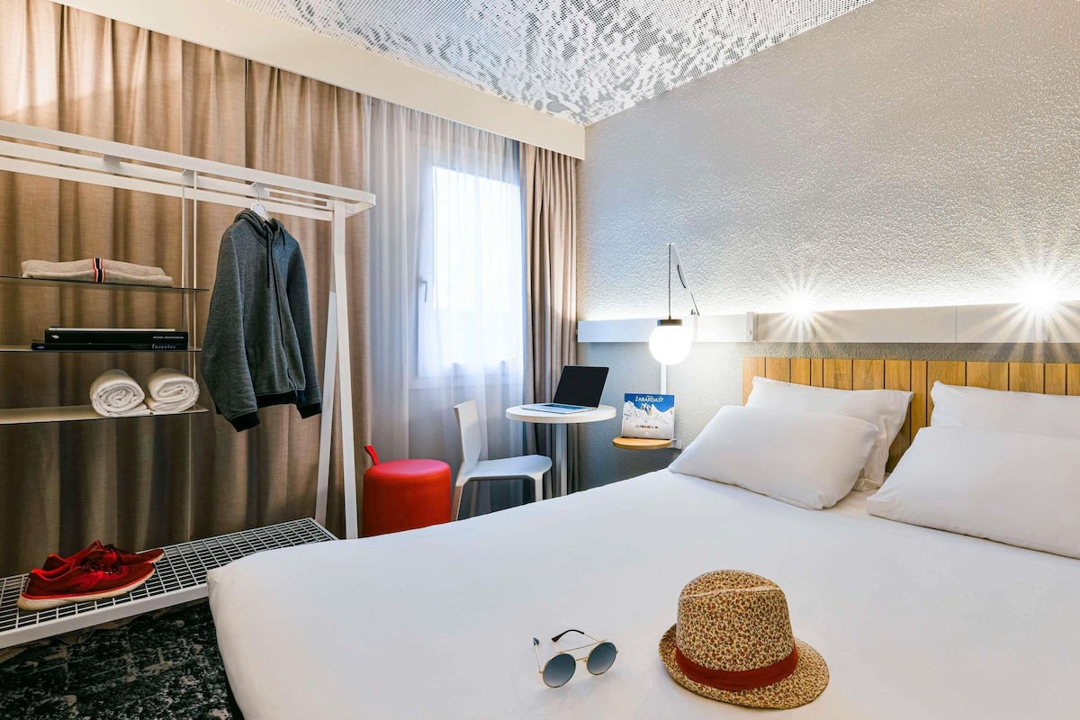 A hotel room with a bed, a desk with a laptop, a red stool, a hat, and a pair of sunglasses on the bed.