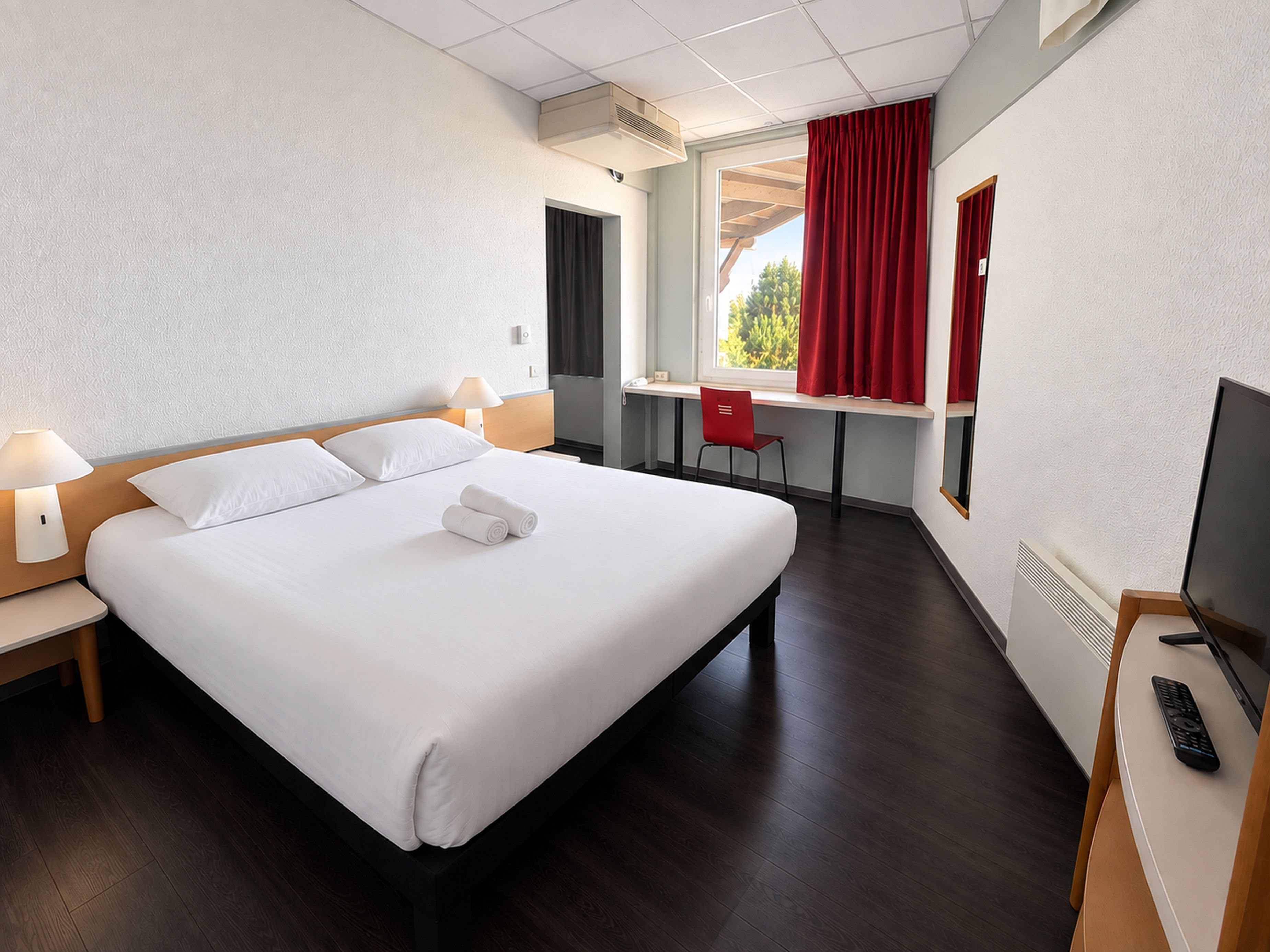Quarto de hotel com cama, escrivaninha, cadeira, televisão e janela com cortinas.