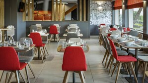 Un restaurante con varias mesas y sillas, algunas con cojines rojos. Las mesas están preparadas con vasos y cubiertos. El interior presenta un diseño moderno con detalles de madera y un gran ventanal en la parte trasera.