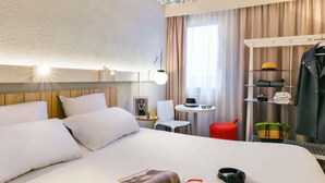 Una habitación de hotel con una cama grande, una mesita de noche con una lámpara, una pequeña mesa con un sombrero y gafas, un reposapiés rojo y un armario con ropa y una cámara.