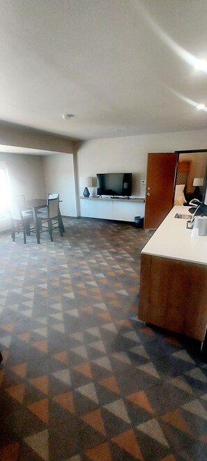 Ein geräumiges Zimmer mit einem gemusterten Teppich, einem Esstisch mit Stühlen, einem Fernseher und einer Küchenzeile.