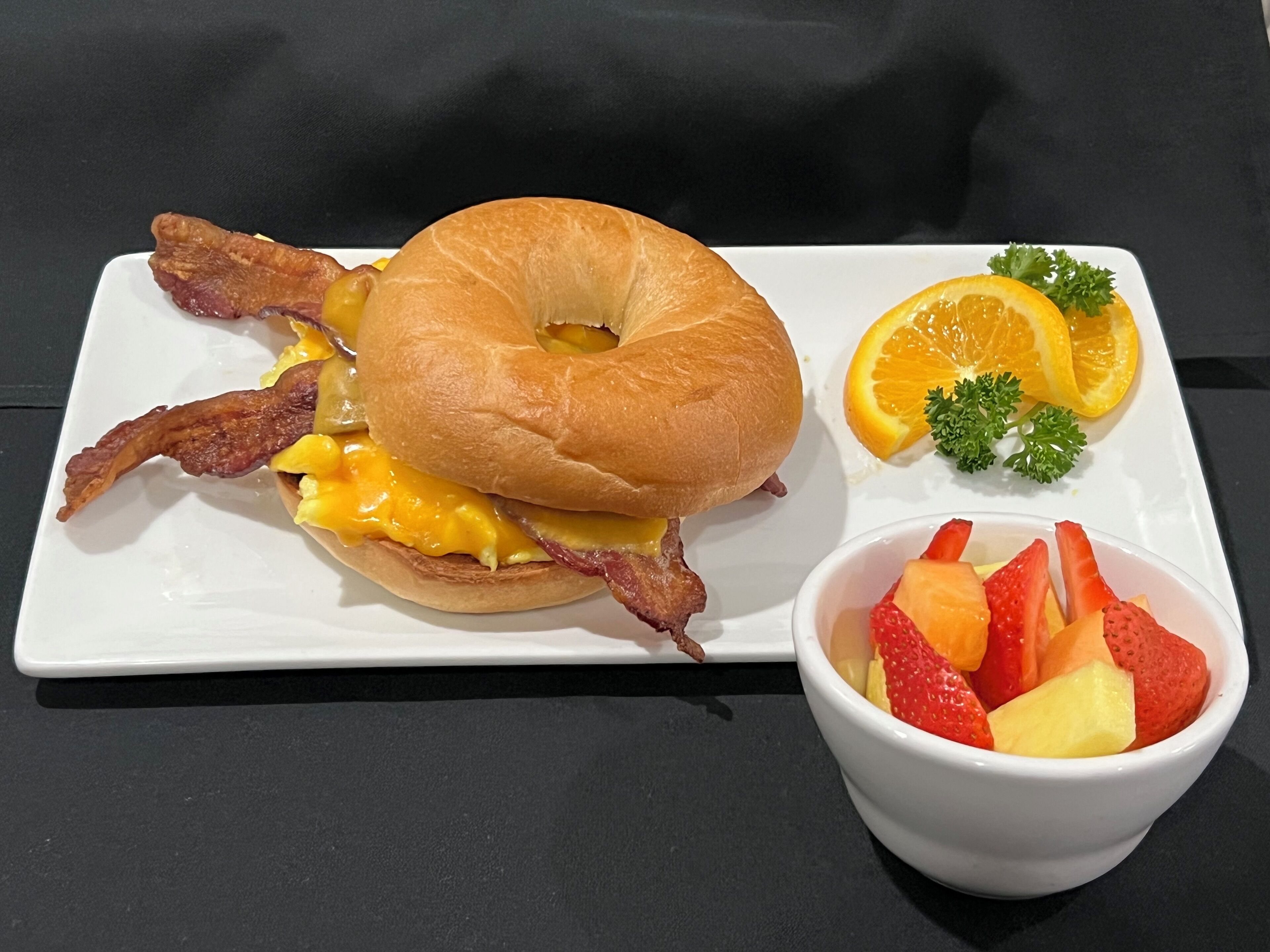A breakfast plate with a bagel sandwich, bacon, cheese, and a fruit bowl containing strawberries, pineapple, and orange slices.