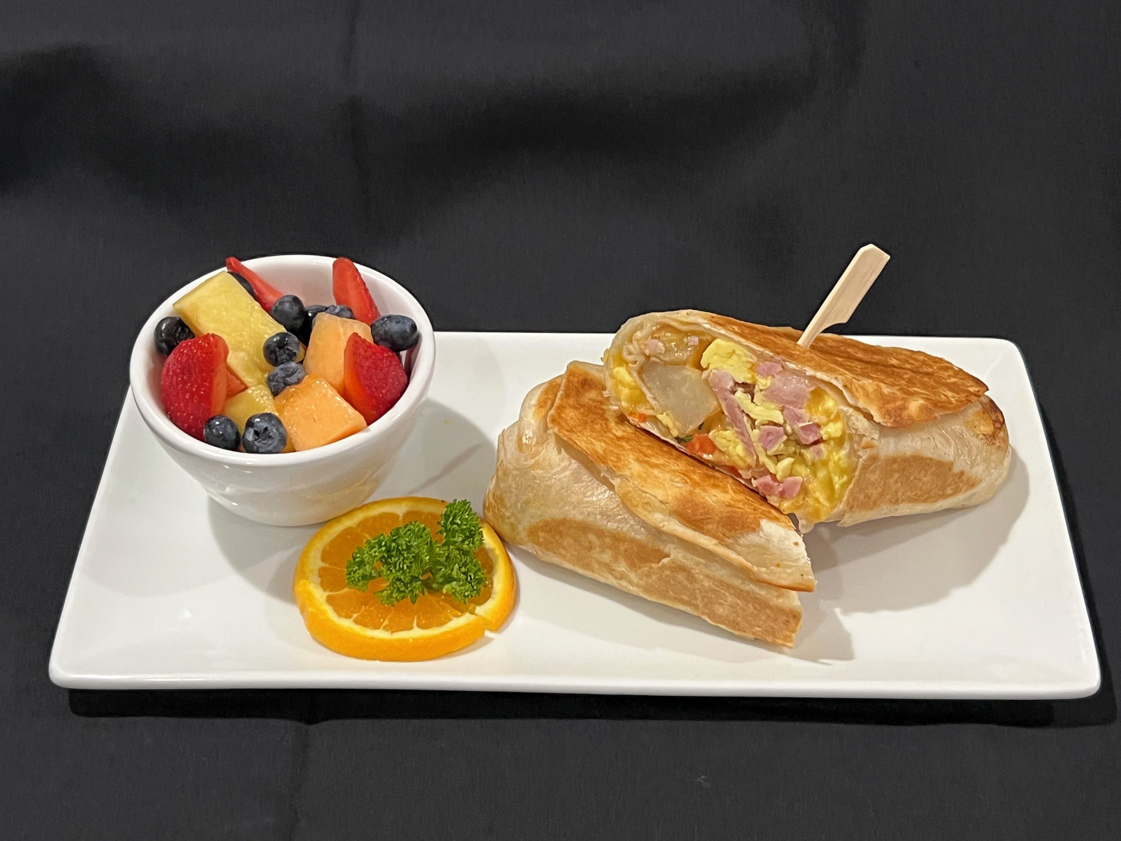 A white plate with a sandwich cut in half, a bowl of mixed fruit, and a slice of orange with parsley garnish.
