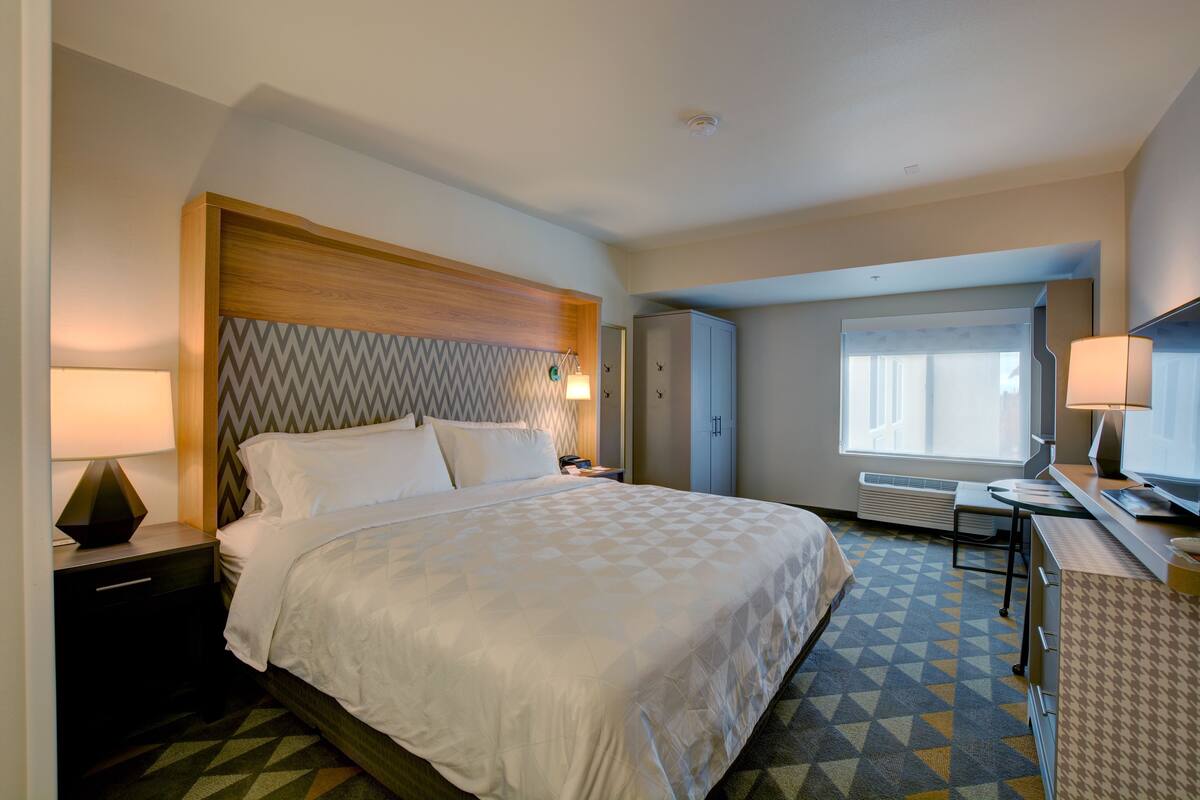 A hotel room with a large bed, a desk, a chair, a lamp, and a window.