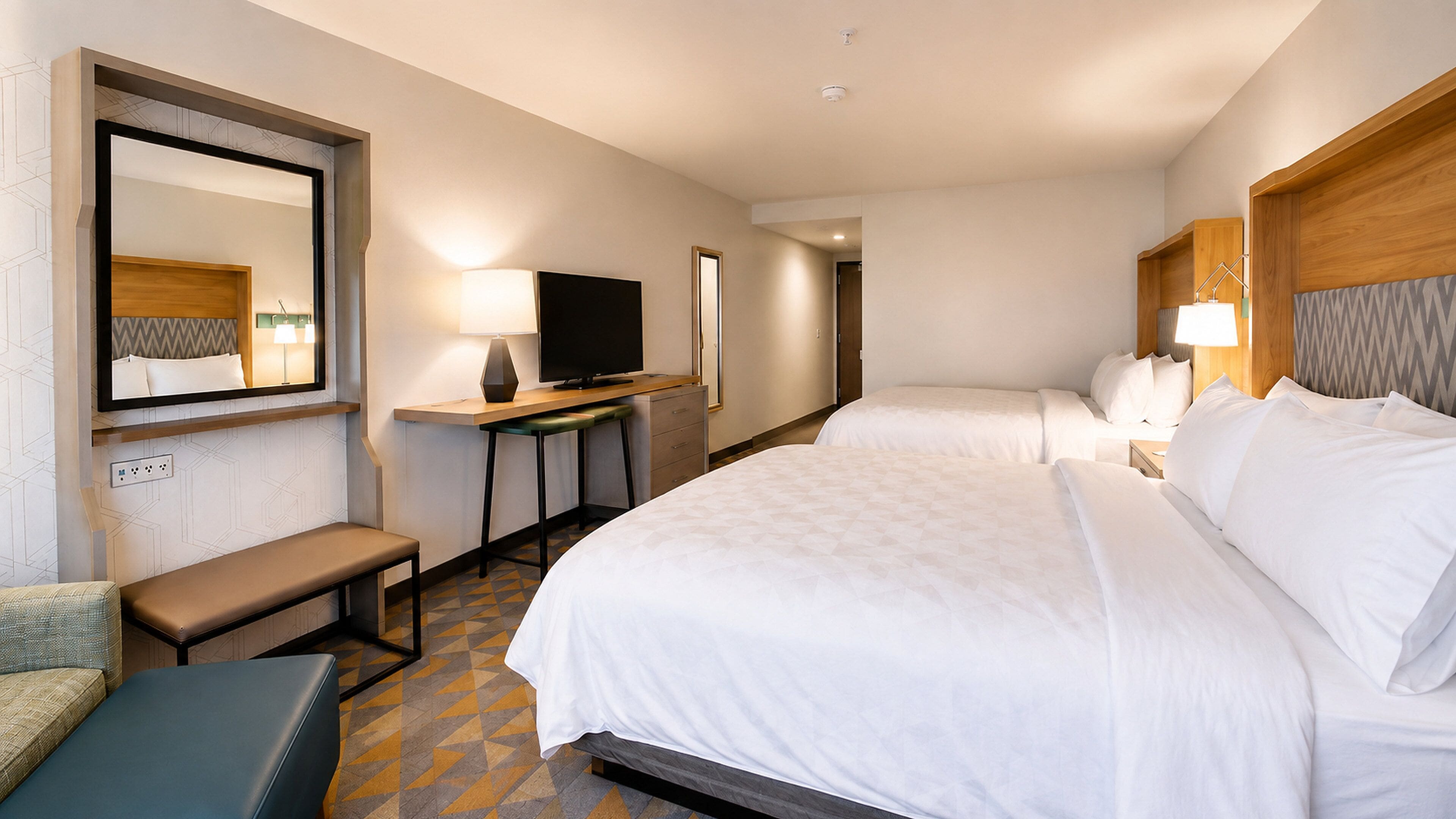 A hotel room with a large bed, a desk with a flat-screen TV, a chair, and a bench.