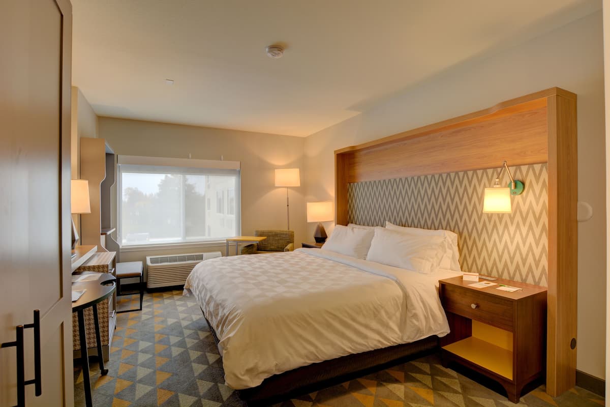 A hotel room with a bed, bedside table, lamp, desk, chair, and a window.