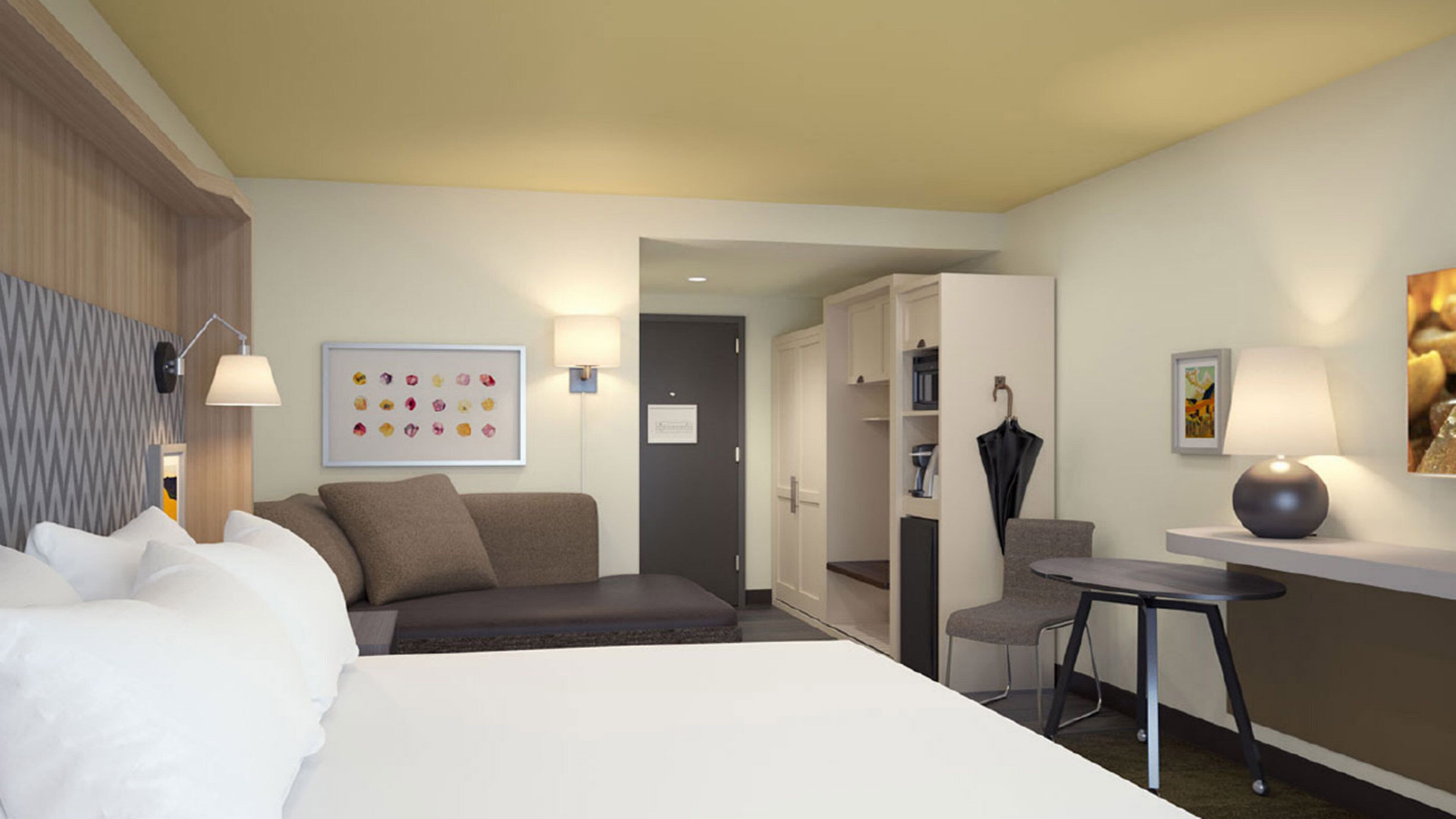 A hotel room with a bed, a sofa, a desk, and a chair. There are wall-mounted lamps, a door, and a framed picture on the wall.