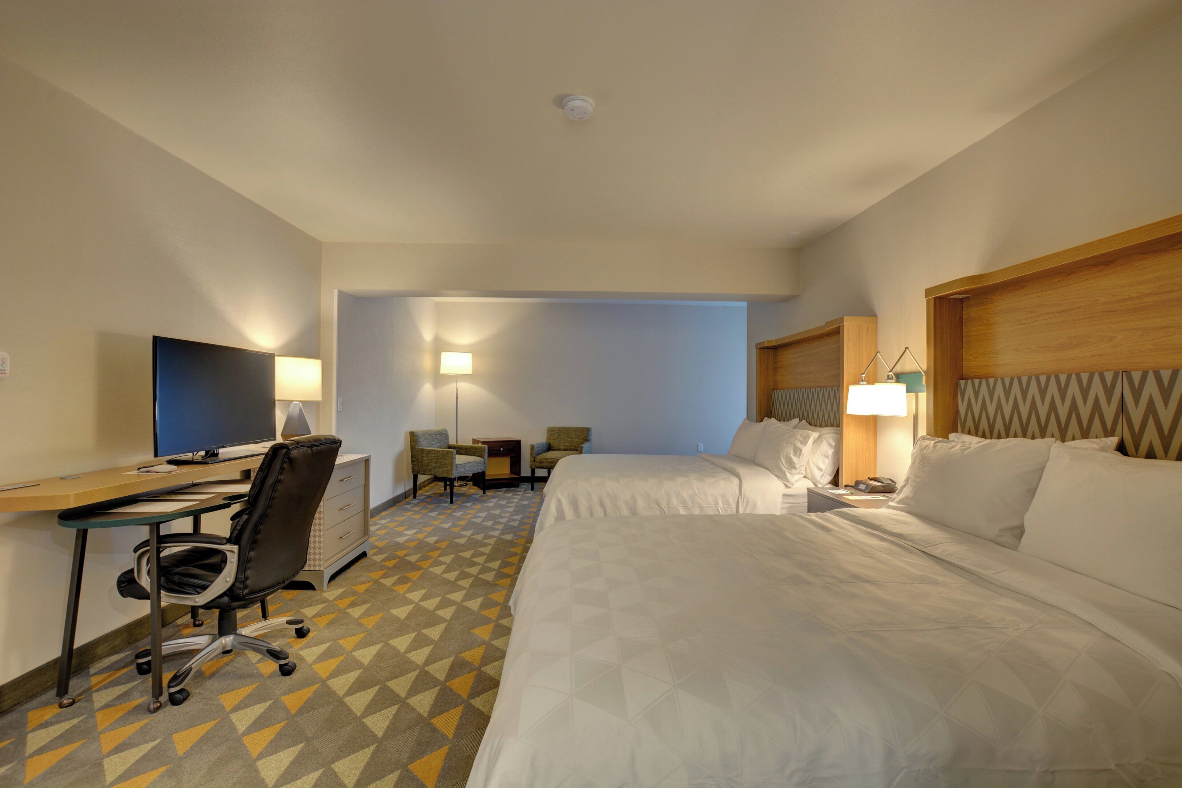 A hotel room with a desk, two chairs, a bed, and a television.