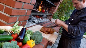 Een bakoven met eten erin, iemand die voedsel op een snijplank bereidt, en diverse groenten op de voorgrond.