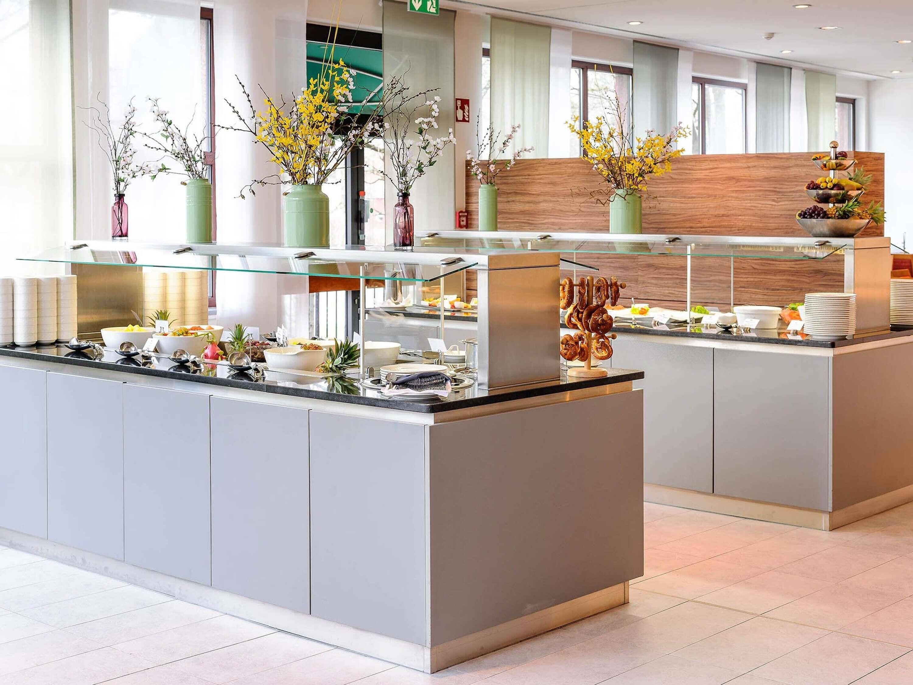 A modern hotel breakfast area with a buffet setup, including various food items and decorative flowers.