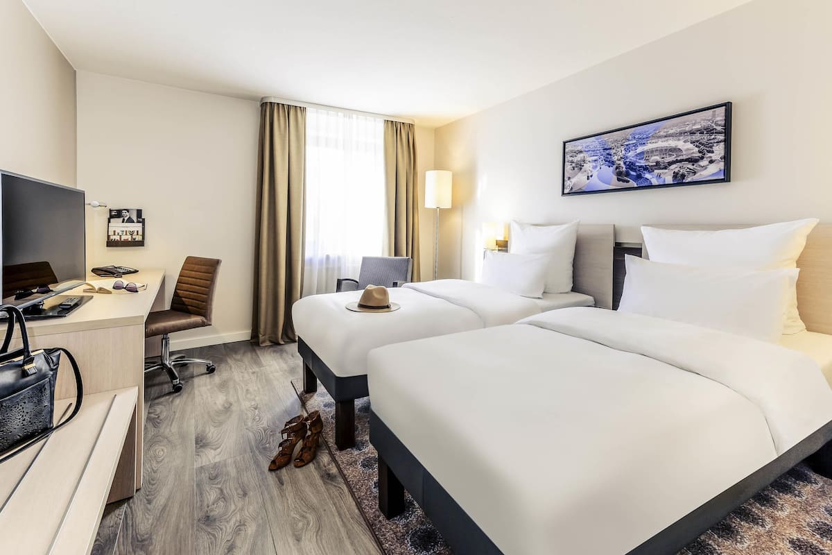 Ein Hotelzimmer mit zwei Betten, einem Schreibtisch mit Computer, einem Stuhl, einem Fernseher und einem gerahmten Bild an der Wand.