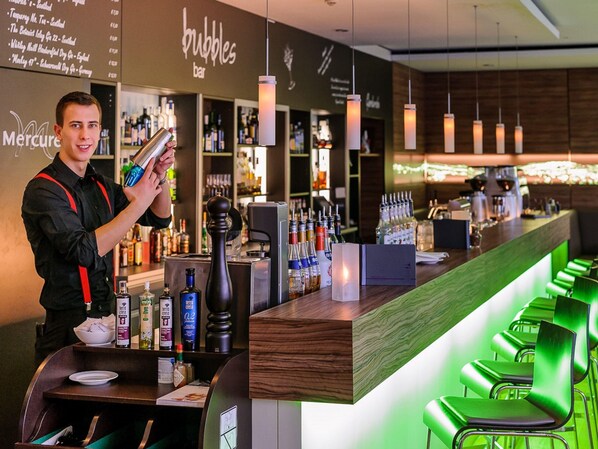 Zona de bar con un cantinero, estantes llenos de botellas de licores y taburetes verdes.