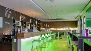 Een moderne bar met een lange toonbank, groene barkrukken en diverse flessen drank.