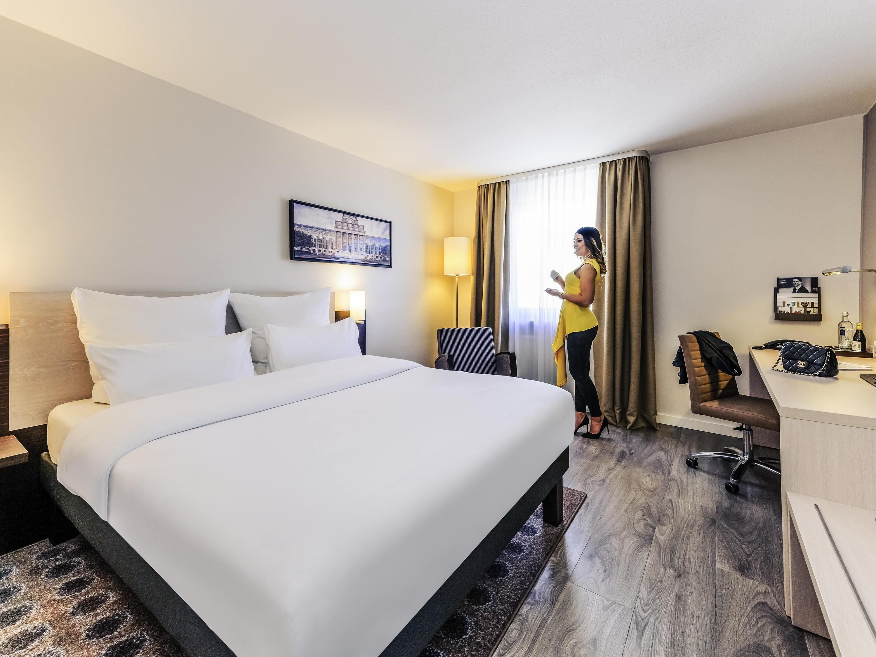 A hotel room with a large bed, a desk, a chair, a lamp, and a window with curtains.
