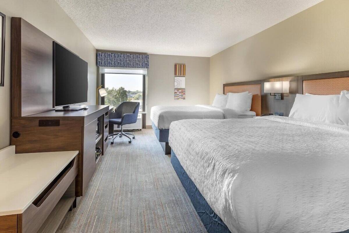 A hotel room with a bed, a desk with a TV, a chair, and a window with a view.