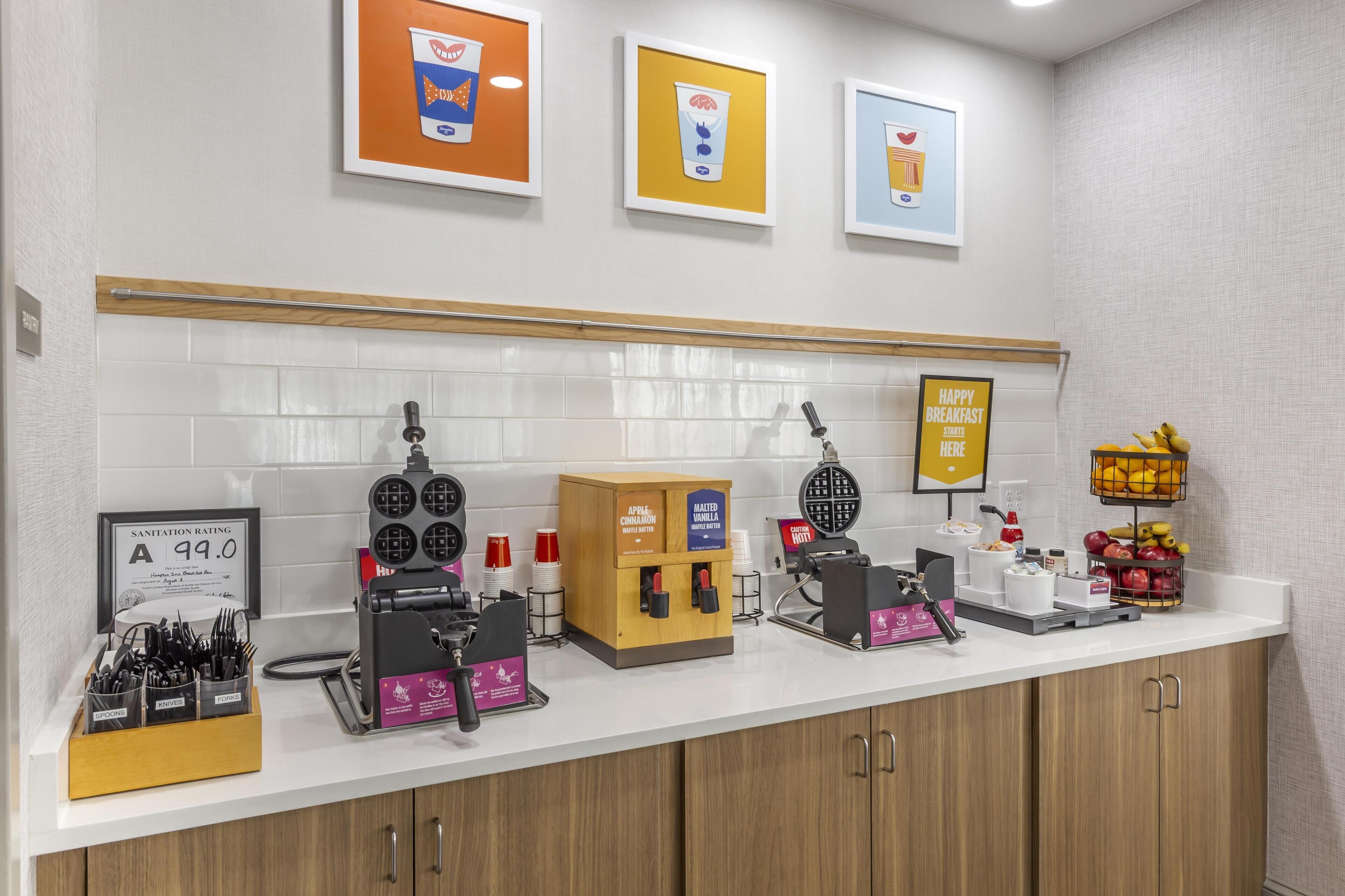 A kitchenette with waffle makers, a sign indicating breakfast starts here, and a fruit basket.