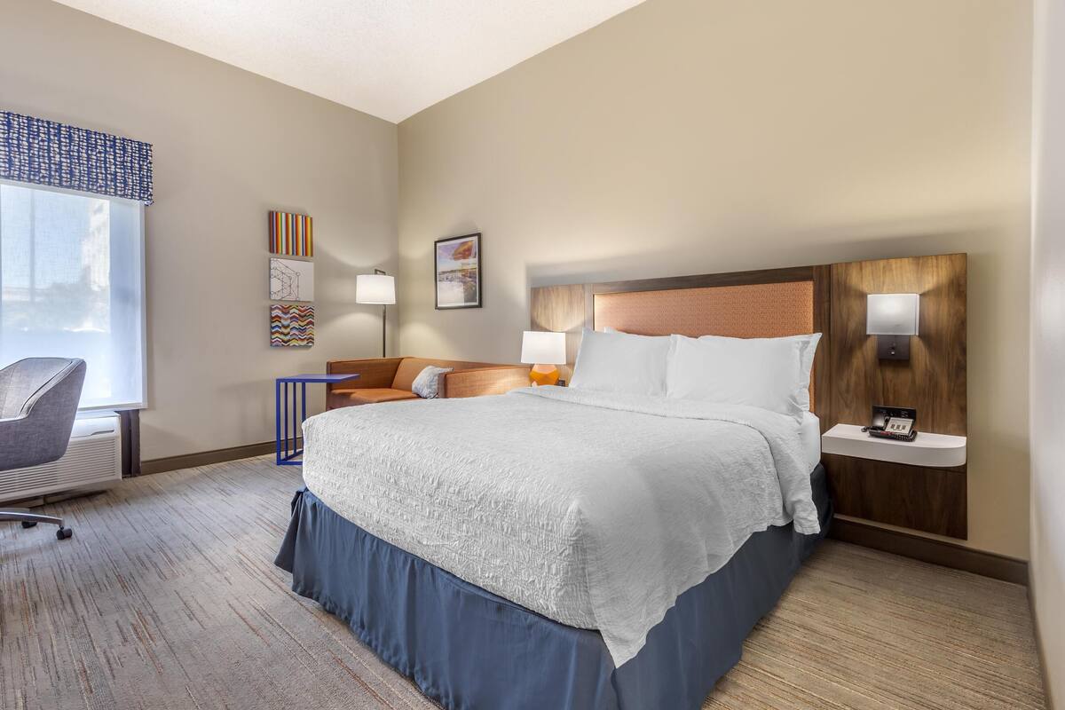 Quarto de hotel com uma cama grande, uma escrivaninha com cadeira, uma mesinha de cabeceira com abajur e obras de arte nas paredes.