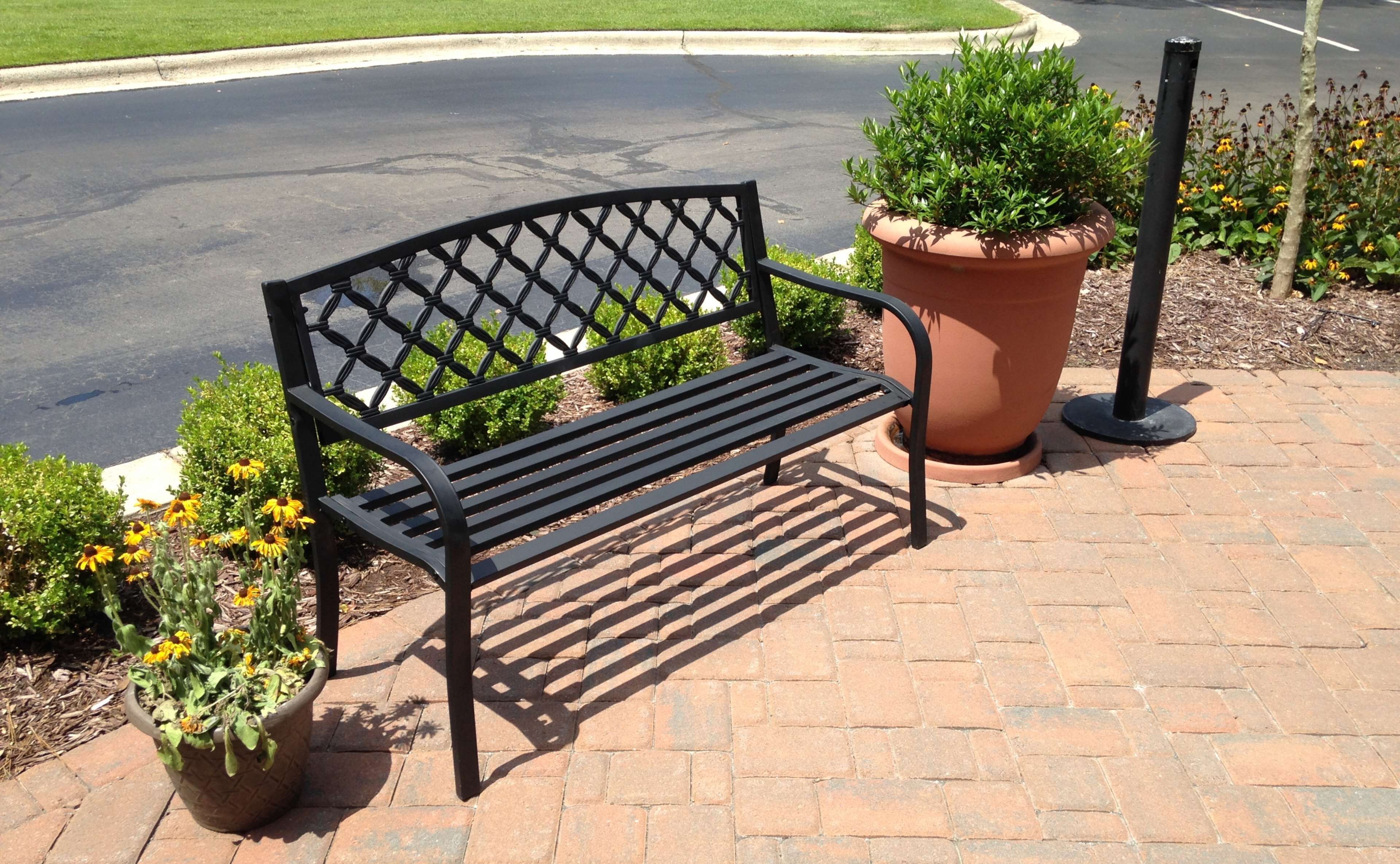A bench with a lattice backrest and armrests, placed on a brick-paved area with potted plants nearby.