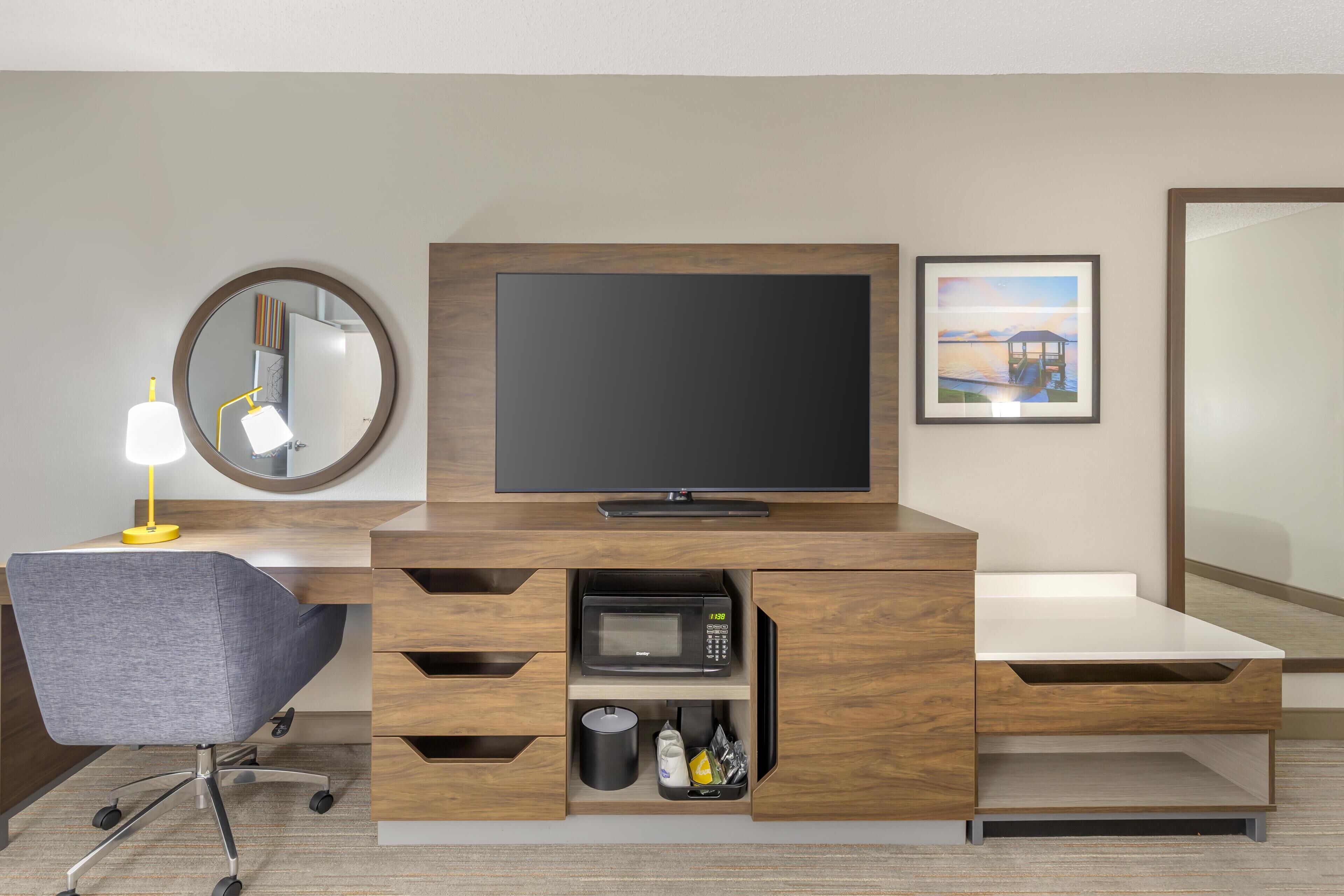 Habitación de hotel con escritorio de madera, televisor de pantalla plana, espejo, cuadro enmarco y microondas.