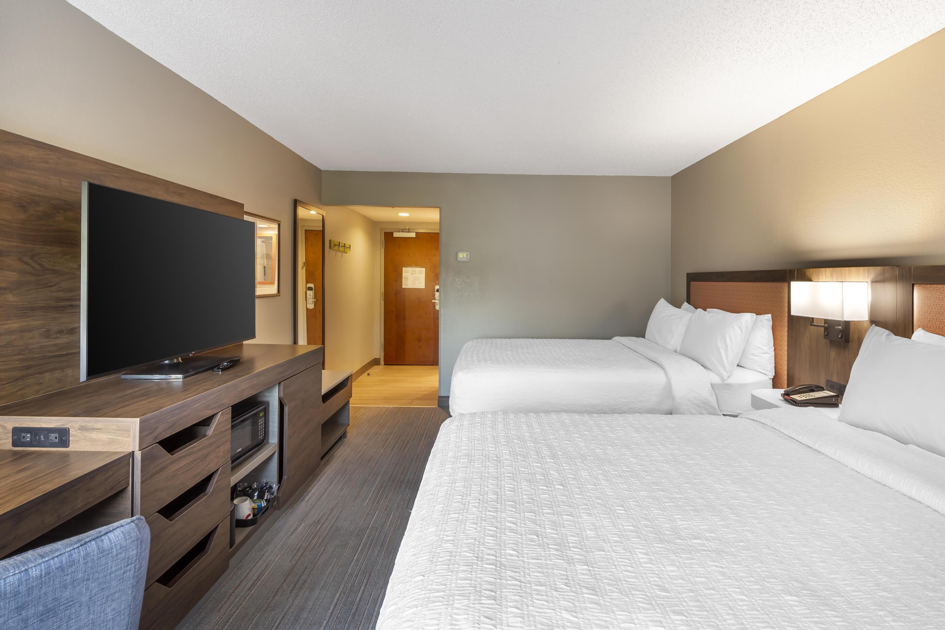 A hotel room with a flat-screen TV, a wooden console table, a microwave, two beds with white bedding, and a nightstand with a lamp.