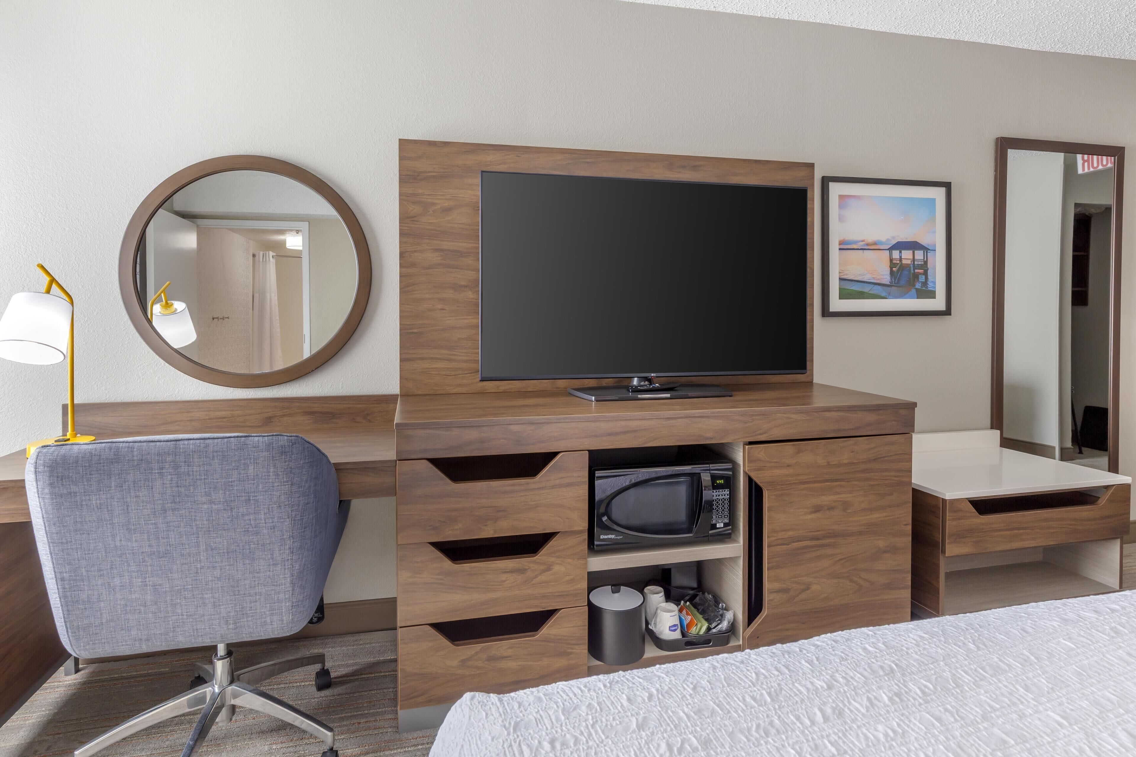 A hotel room with a wooden desk, a flat-screen TV, a microwave, a chair, and a bed.