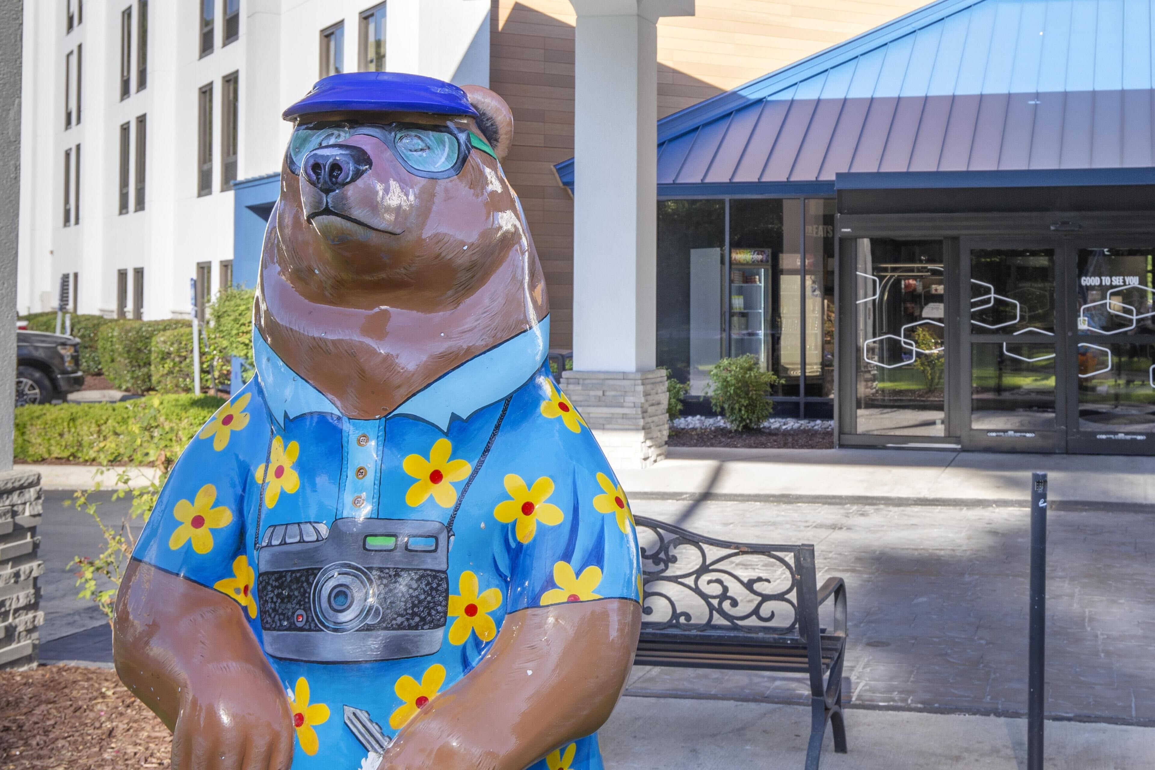 Une statue d’un ours portant une chemise bleue ornée de fleurs jaunes, qui se trouve devant un bâtiment doté d’un toit bleu et près d’un banc.