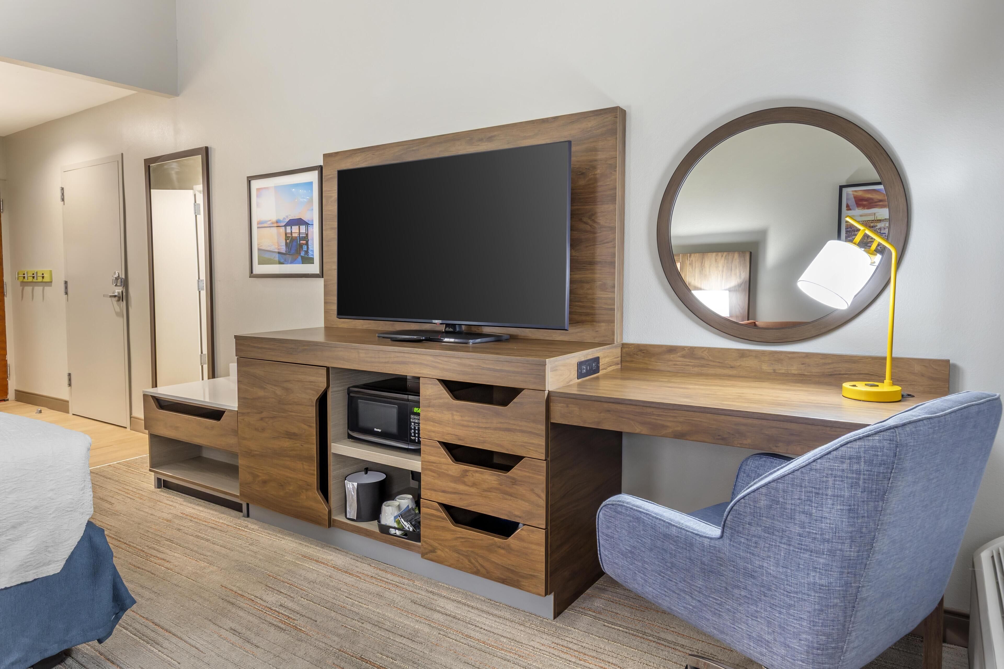 Habitación de hotel moderna con un escritorio de madera, un televisor de pantalla plana, un espejo redondo y una silla azul.