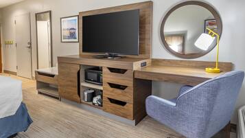 A modern hotel room with a wooden desk, a flat-screen TV, a round mirror, and a blue chair.