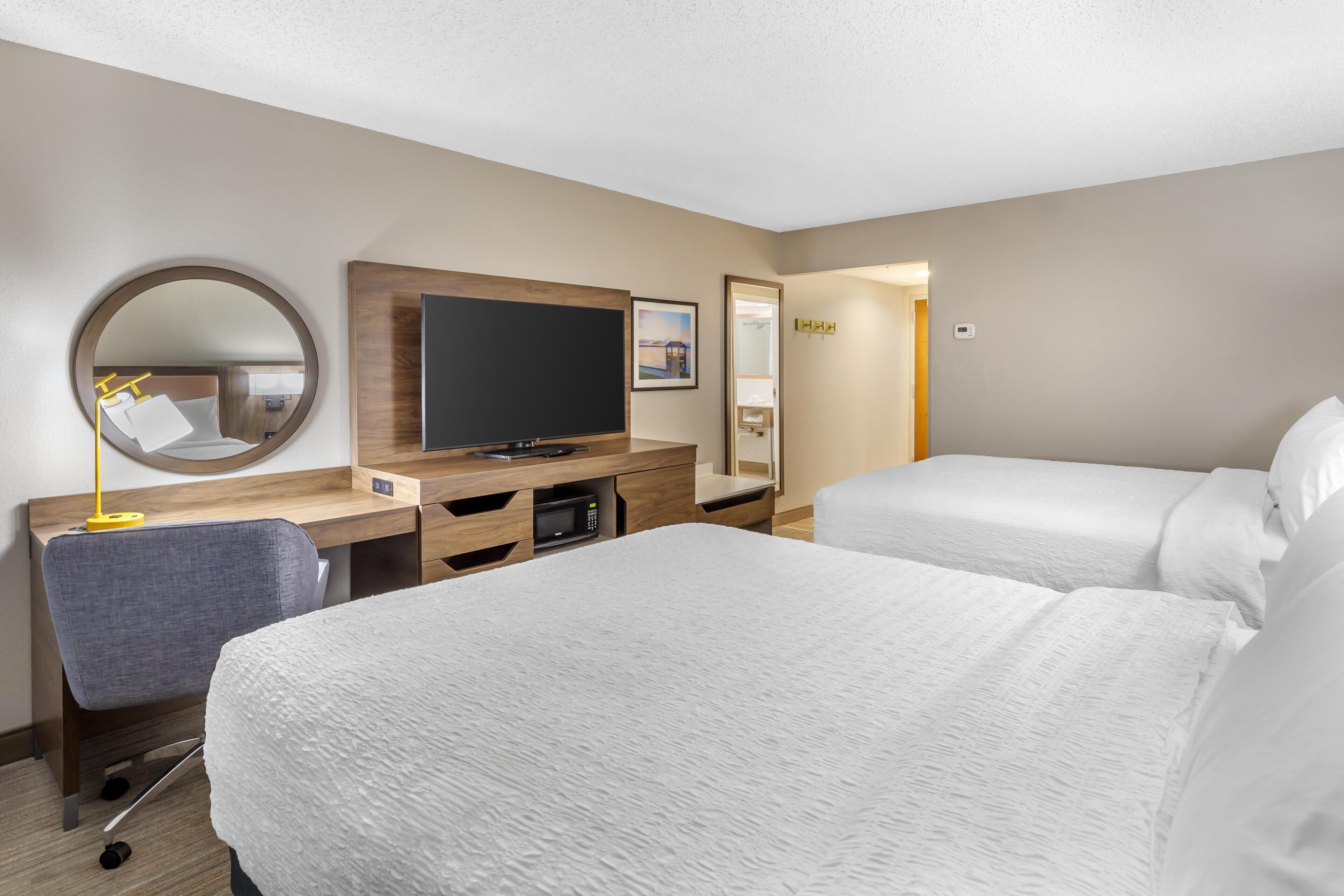 Quarto de hotel com duas camas, uma escrivaninha, televisão de tela plana, espelho e um pequeno espaço para sentar.