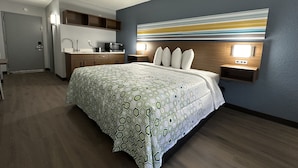 A bedroom with a bed, a nightstand, a wall-mounted TV, and a kitchenette area. - HomeTowne Studios by Red Roof Richmond, VA - W Broad St (Richmond)