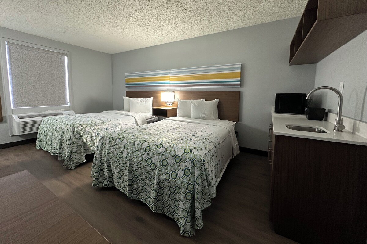 A hotel room with two beds, a large window, a modern bathroom, and a decorative wall piece.